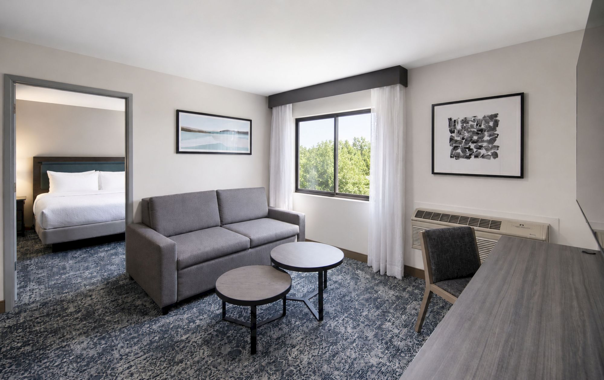 A modern hotel living room with a gray sofa, round tables, large window with curtains, framed art, and an air conditioner.