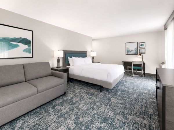 A modern hotel suite with a bed, gray sofa, desk, framed art, and blue patterned carpet&mdash;bright and tidy.