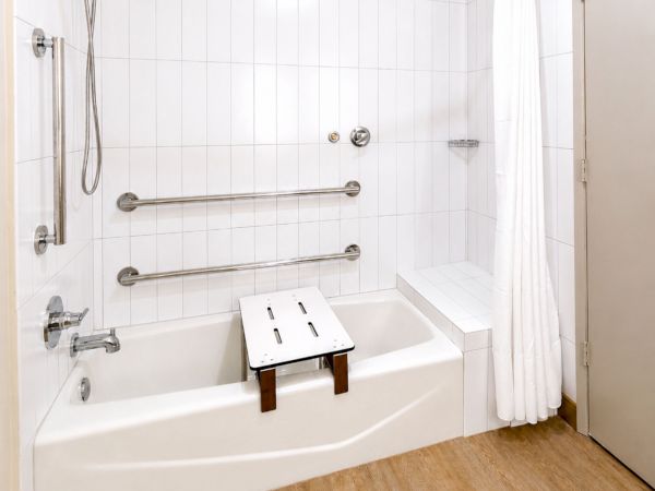 A bright accessible bathroom with a bathtub, grab bars, a showerhead, and a small transfer bench with a safety shower curtain.