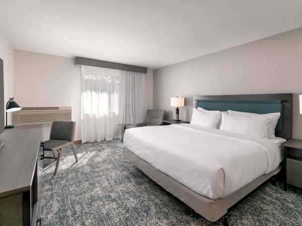 A modern hotel room with a king bed, bedside lamps, desk area, and sheer curtains.