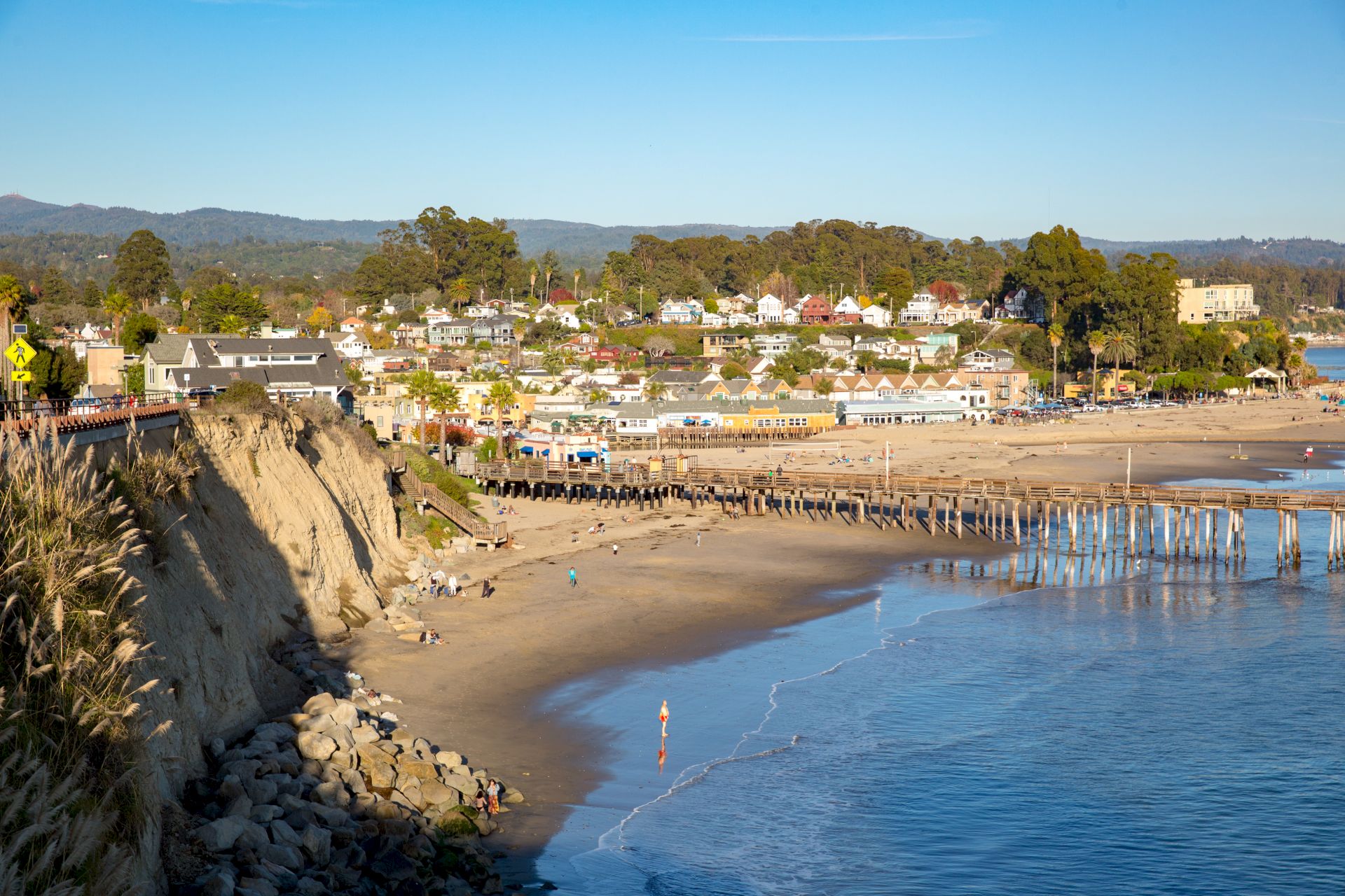 Sunny seaside town with a sandy beach, rocky shoreline, and a long wooden pier stretching into blue water.