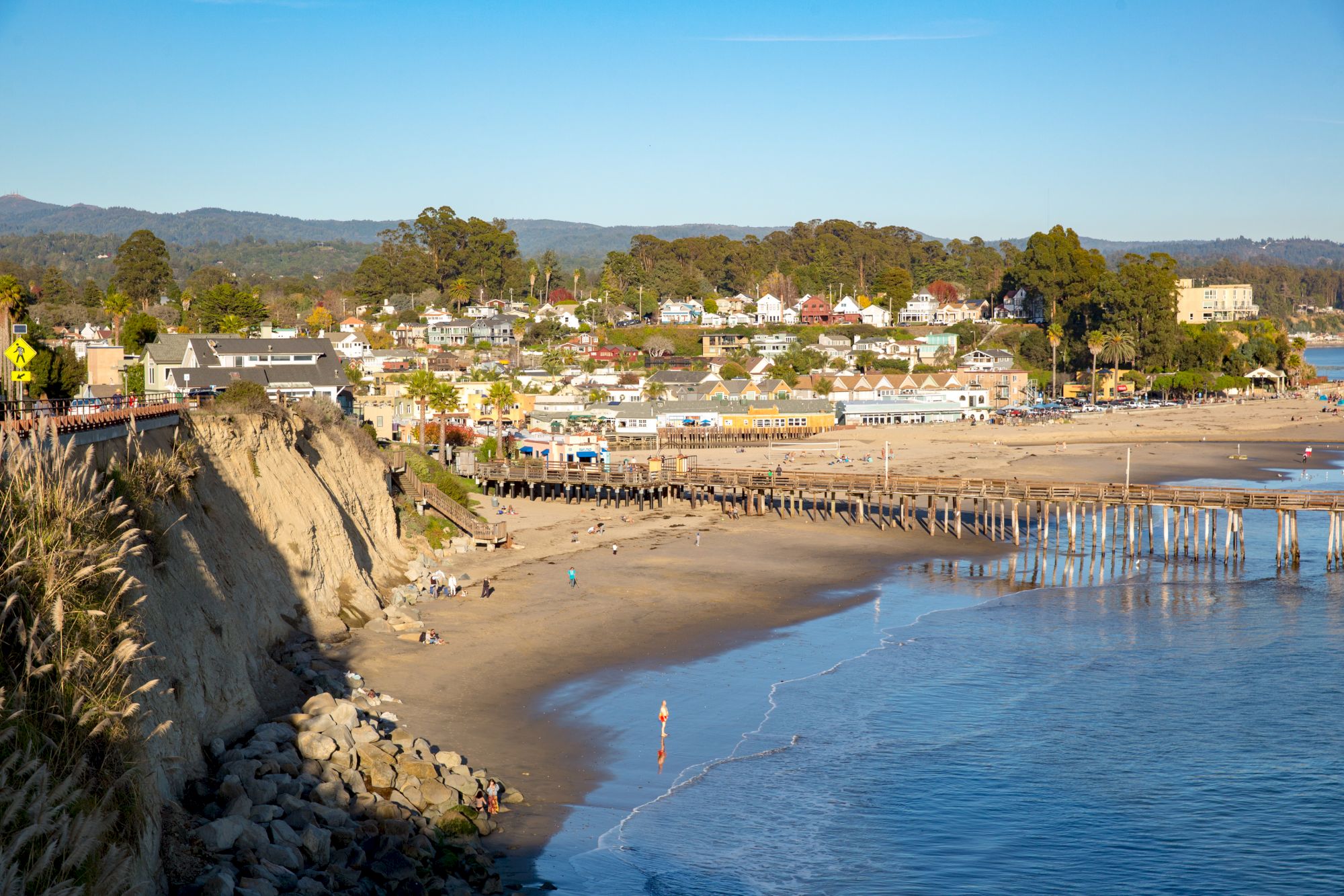 Sunny seaside town with a sandy beach, rocky shoreline, and a long wooden pier stretching into blue water.