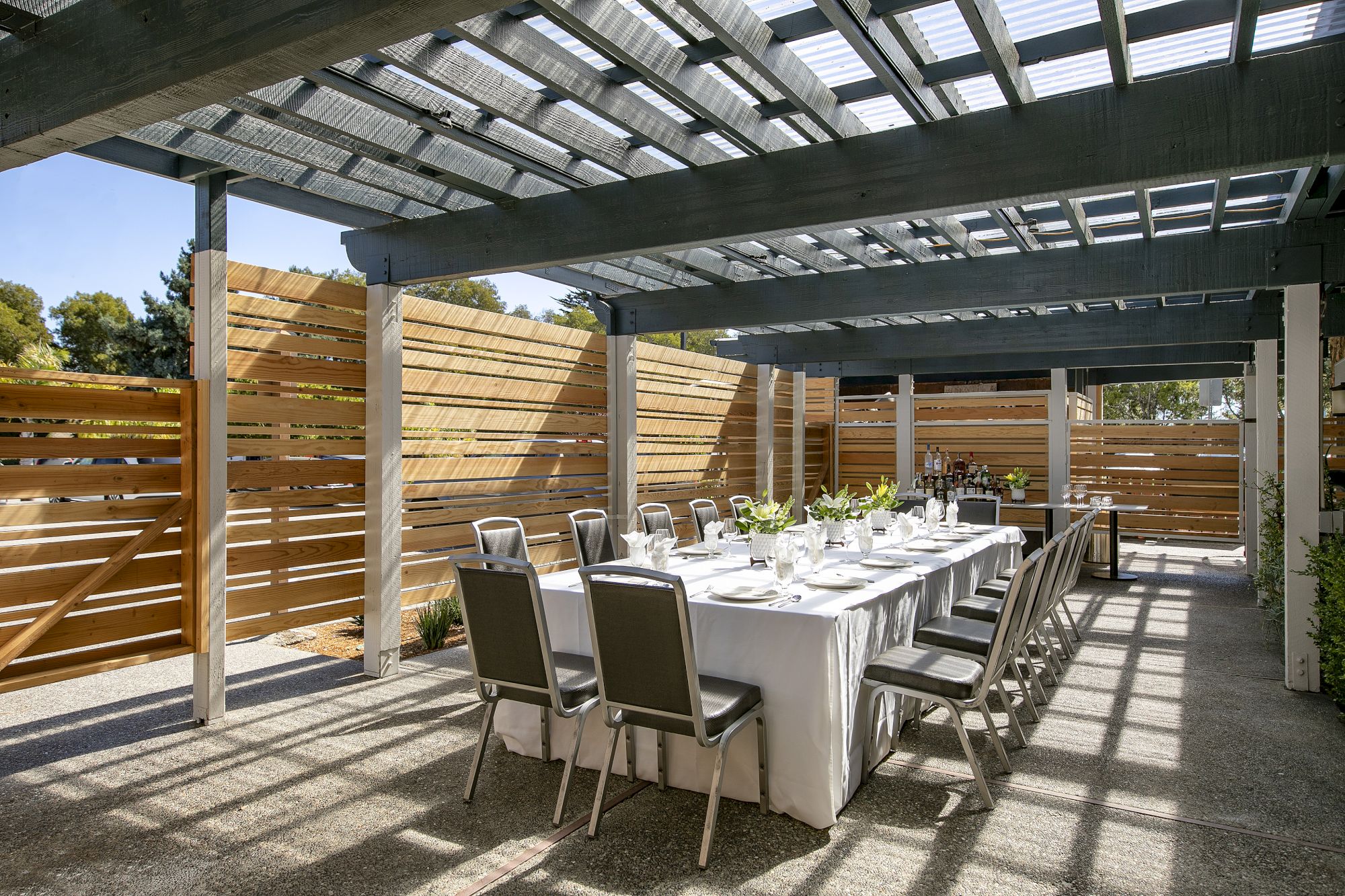 An outdoor patio dining setup with a long table, chairs, and a wooden pergola sheltering bottles and flowers.