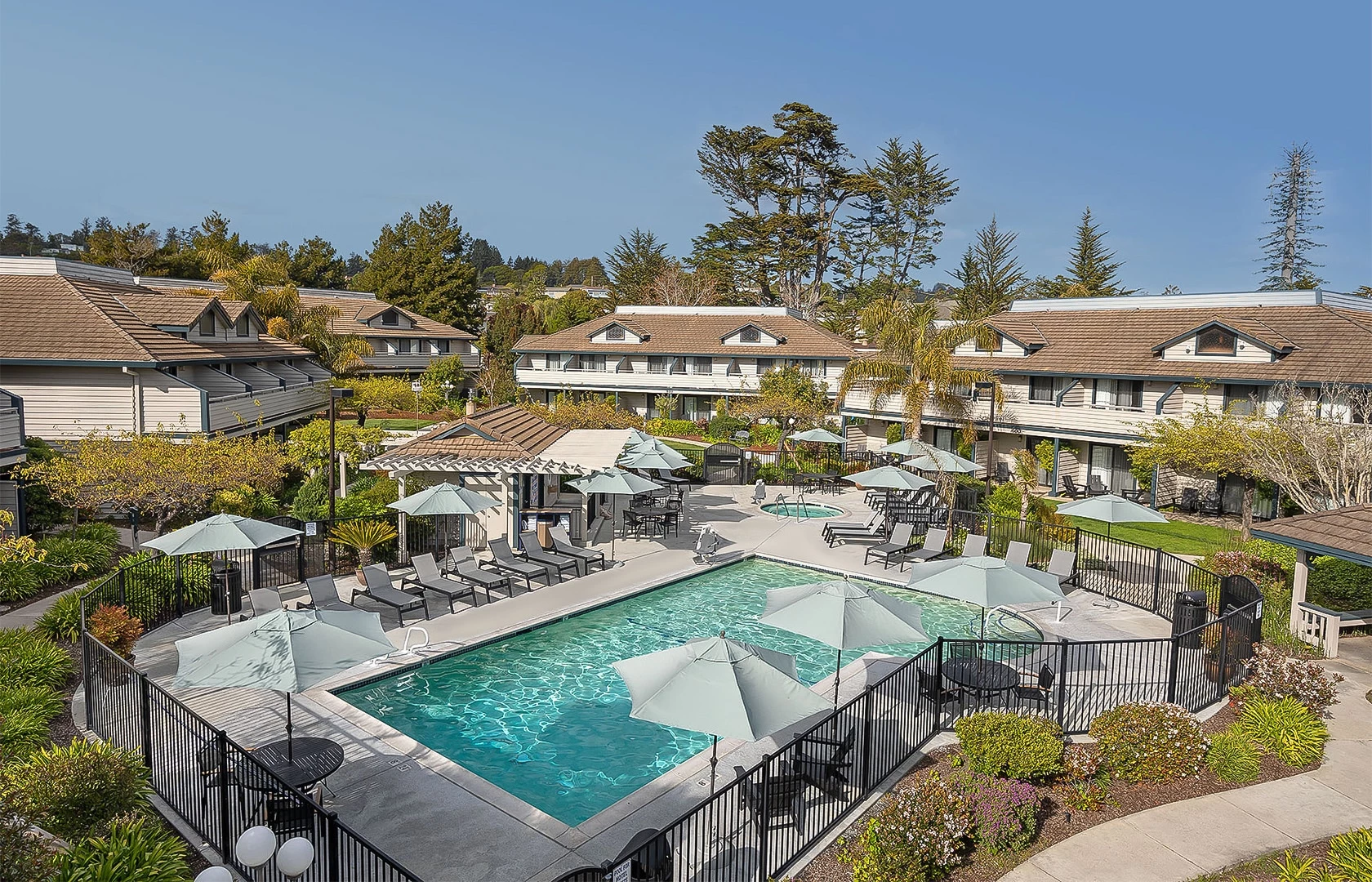 A sunny pool area with a fenced rectangular pool, lounge chairs under umbrellas, and tan multi-unit buildings around the courtyard.