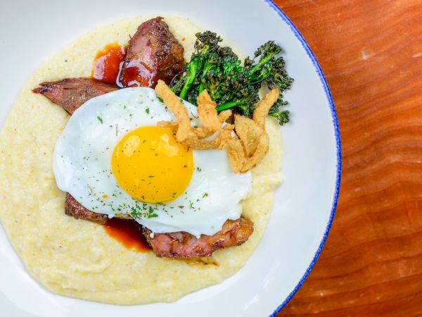 A plate of creamy polenta topped with a sunny-side-up egg, bacon, crispy bits, and broccoli on the side.