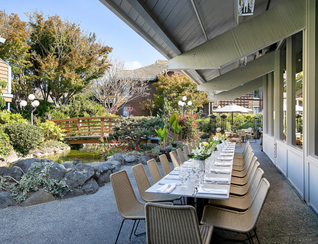 An elegant outdoor patio set for a meal, with neatly placed chairs, tables, flowers, and a small garden pond beside it.