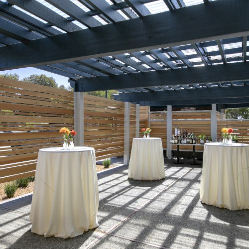 An outdoor patio with a wooden pergola, white-covered tables, bottles and flowers, and sunlit gravel under dark beams.