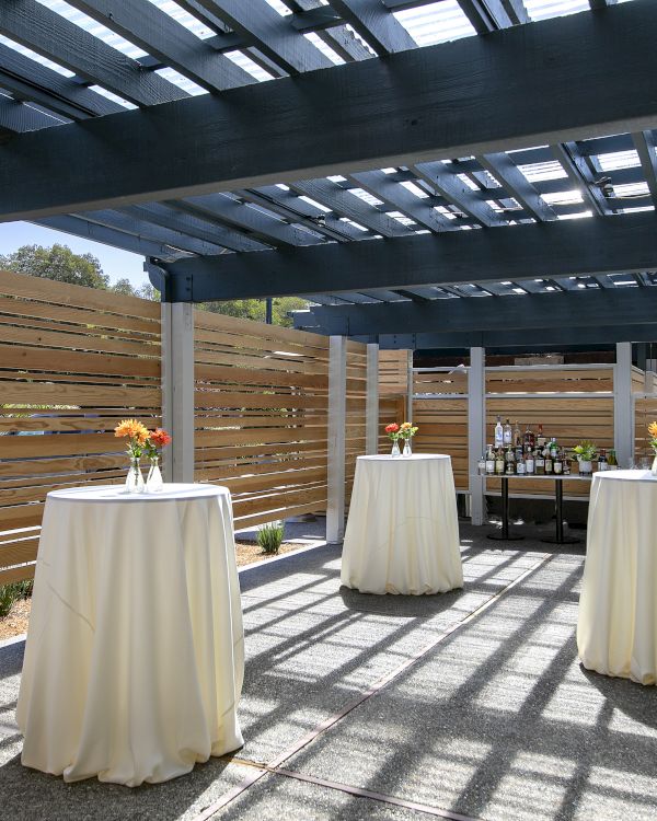 An outdoor patio with a wooden pergola, white-covered tables, bottles and flowers, and sunlit gravel under dark beams.