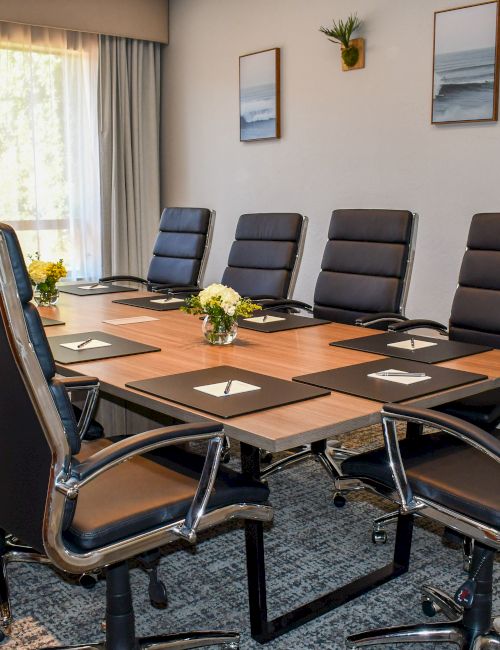 A modern conference room with a long wooden table, black leather chairs, pens and notepads, and framed ocean artwork.