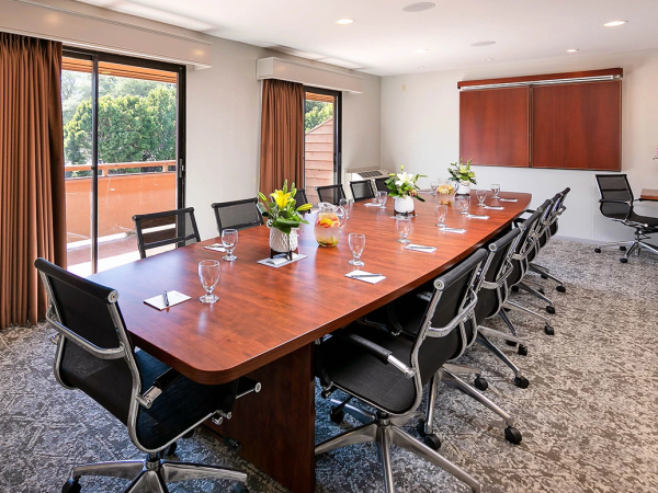 A modern boardroom with a long wooden table, black office chairs, glassware and flowers, large windows with brown curtains, and a clean, professional setup.