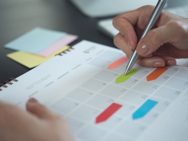 A person uses a pen to mark a monthly calendar, reviewing colorful schedule blocks and numbers.