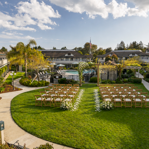 A manicured resort garden with chairs set for an outdoor ceremony, a central aisle, and a pool backdrop under a blue sky.