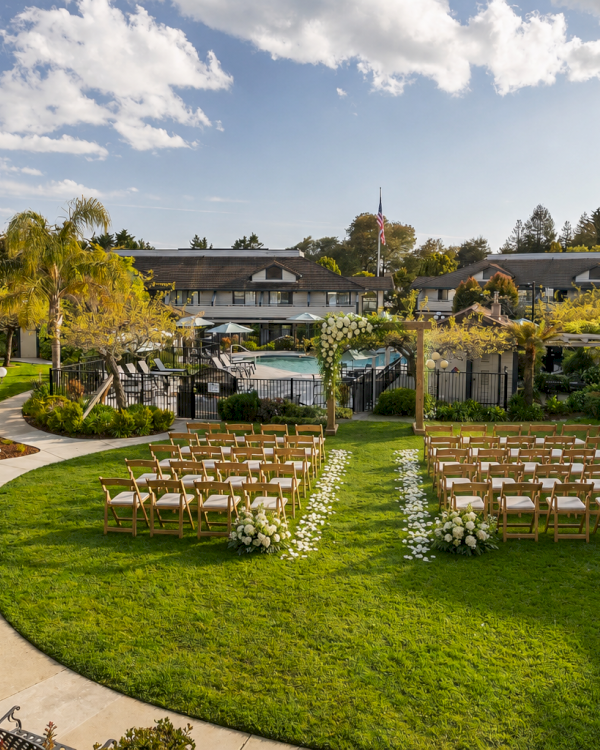A manicured resort garden with chairs set for an outdoor ceremony, a central aisle, and a pool backdrop under a blue sky.