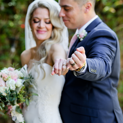 A bride and groom outdoors in wedding attire, smiling softly while touching hands with rings on their fingers.
