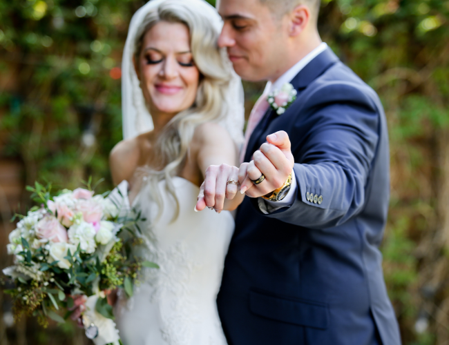 A bride and groom outdoors in wedding attire, smiling softly while touching hands with rings on their fingers.