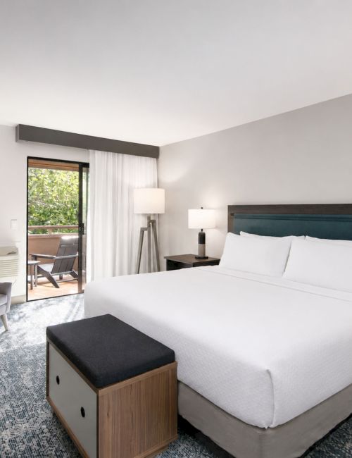 A modern hotel room with a king bed, nightstands, lamps, armchair, framed art, and a sliding door to a patio.