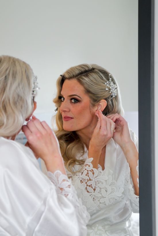 A blonde woman sits at a mirror adjusting her hair and jewelry while another person gently applies makeup in a bright room.