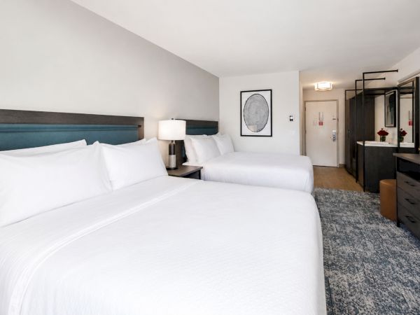 A modern hotel room with two white beds, bedside lamps, a framed print, and a dresser with a mirror.