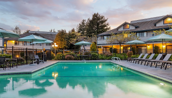 An outdoor swimming pool at sunset with lounge chairs, umbrellas, and townhouses in the background.