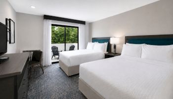 A modern hotel room with two white beds, teal headboards, a desk with TV, and a large window showing lush greenery.