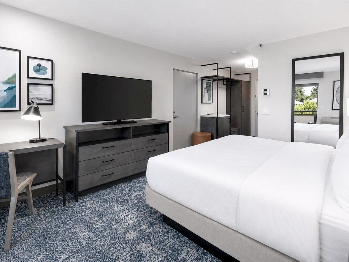 A modern hotel-style bedroom with a white bed, gray dresser, flat-screen TV, lamp, desk, and framed wall art.