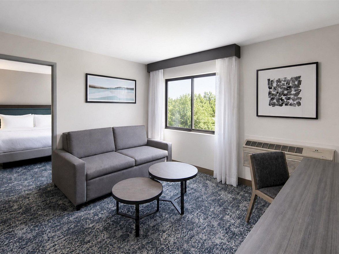 A modern hotel living area with a gray sofa, round tables, large window, framed art, and an AC unit by the desk.