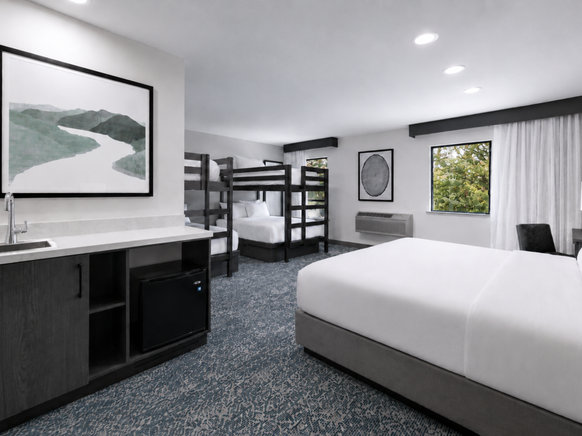 Modern hotel room with two beds, a bunk set, a large vanity, framed art, curtains, and windows overlooking greenery.