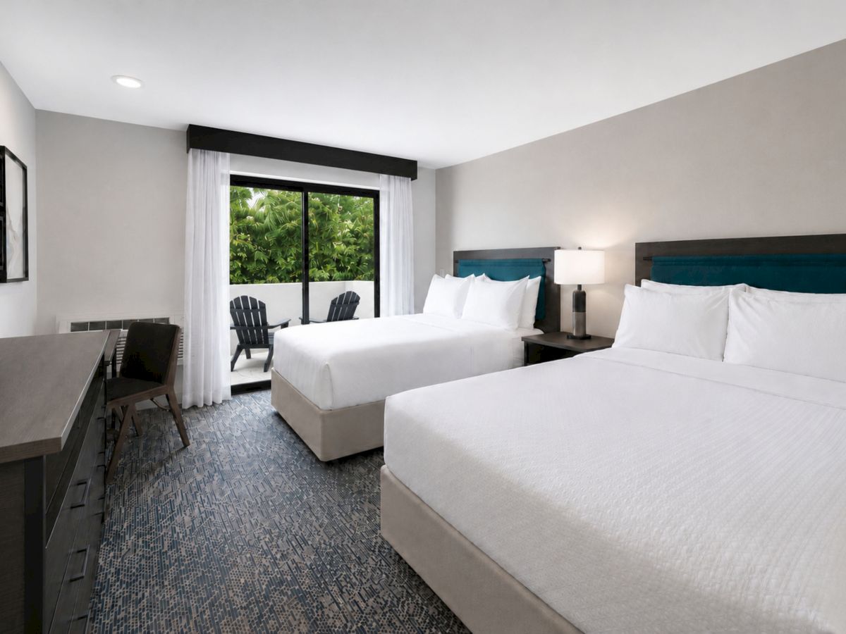 A modern hotel room with two white beds, teal headboards, a desk, and a large window with outdoor greenery view.