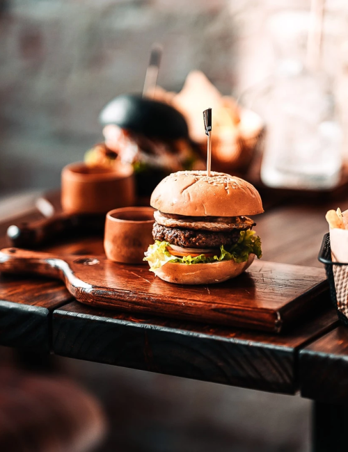 A chef&rsquo;s counter with a burger and iced cola in a glass, ready to grab with fries and sauce.