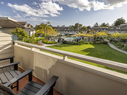 A sunny apartment courtyard with manicured green lawn, trees with yellow blossoms, black fences, and a distant pool area under a blue sky.