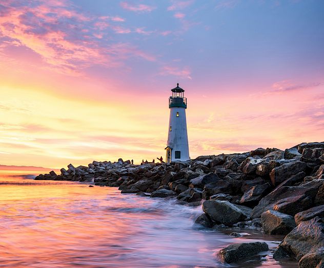 A white lighthouse stands on a rocky shoreline at sunset, with waves rolling in and pink clouds lighting the sky.