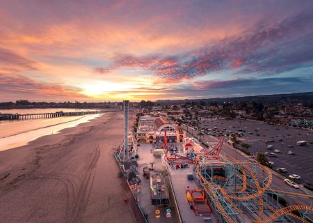 Aerial view of a coastal amusement pier by the beach at sunset, with colorful roller-coaster tracks and docks along the shore.