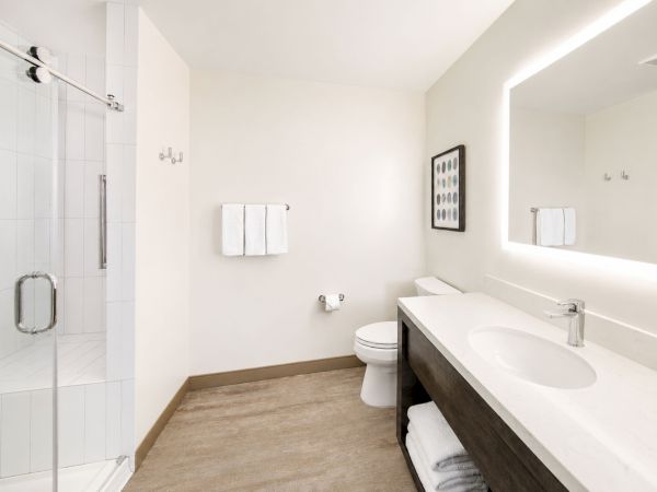 Bright modern bathroom with a glass shower, white tiles, vanity sink, toilet, towel bars, and framed artwork.