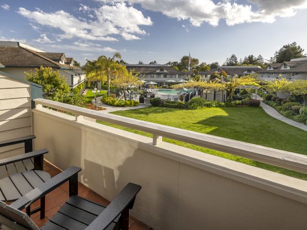 View of a sunny residential courtyard with manicured grass, trees, fencing, and a pool between apartment buildings.