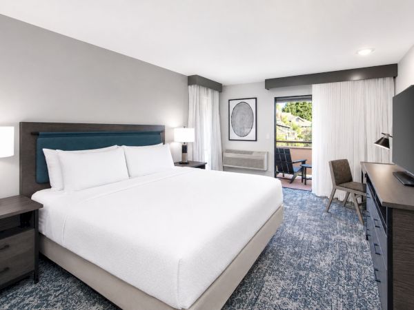 A modern hotel room with a large bed, nightstands, a desk and chair, a wall TV, curtains, and window view.