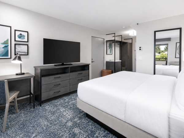 A modern hotel room with a white bed, flat-screen TV, dresser, framed art, and dark wood accents under bright lighting.