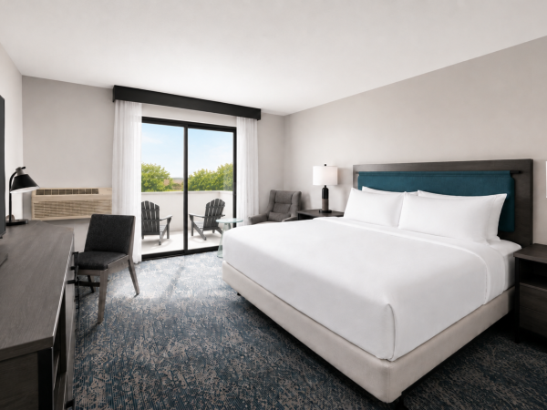 Modern hotel room with a white bed, bedside lamps, seating by a sliding door window, and outdoor greenery.