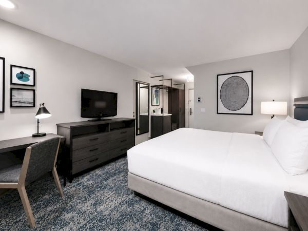 A modern hotel room with a white bed, dresser/TV setup, framed art, and sliding glass doors in the background.