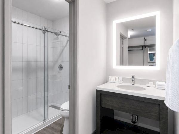 Bright modern bathroom with glass shower, tiled walls, toilet, and dark vanity sink under a lit mirror.