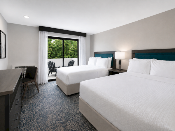 A modern hotel room with two neatly made beds, bedside lamps, and large windows/door overlooking green foliage.
