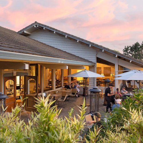 A cozy outdoor restaurant terrace at sunset with people dining under umbrellas and warm lights along a landscaped garden path.
