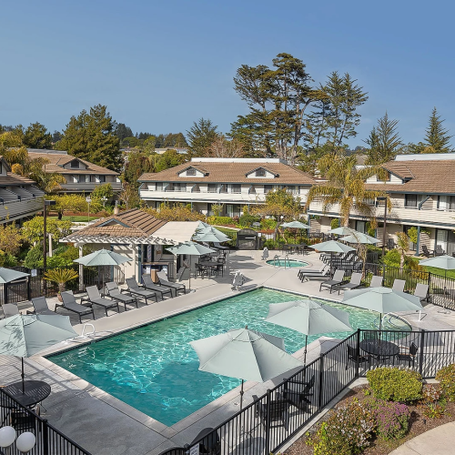 A hotel courtyard with a turquoise swimming pool, sun umbrellas, lounge chairs, and surrounding townhouses under a blue sky.