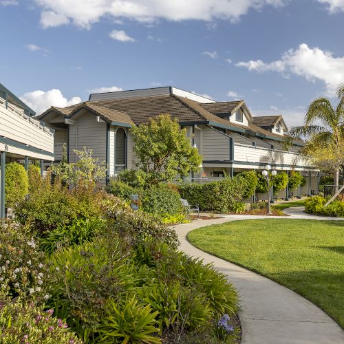 A modern apartment complex with beige buildings, green railings, manicured hedges, palm trees, and a sunny blue sky.