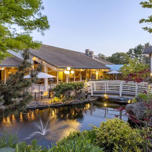 A tranquil resort courtyard with a pond and fountain, warm-lit buildings, and lush trees under a blue evening sky.
