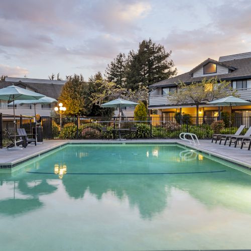 A quiet hotel courtyard pool at dusk with green umbrellas, lounge chairs, and lit buildings under a colorful sky.