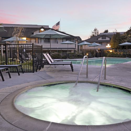 A backyard resort-style pool and hot tub at dusk, with lounge chairs, umbrellas, a fence, and a small U.S. flag by the buildings.