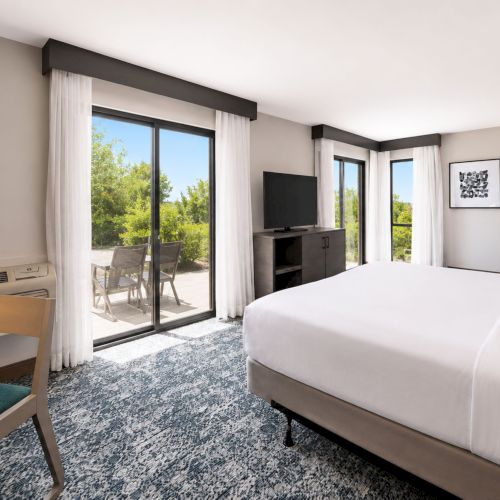 A modern hotel bedroom with a white bed, flat-screen TV, dark beams, sheer curtains, and floor-to-ceiling doors opening to a patio.