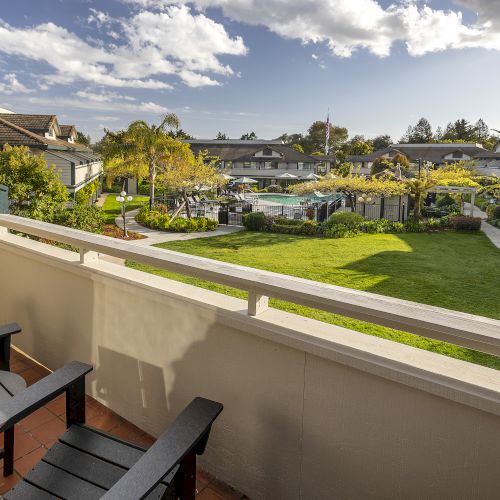 A sunny residential courtyard with a lawn, trees and a fenced pool, viewed from a balcony with two chairs.
