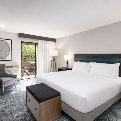A modern hotel bedroom with a large bed, bedside lamps, armchair, framed art, and an open sliding door to a balcony.