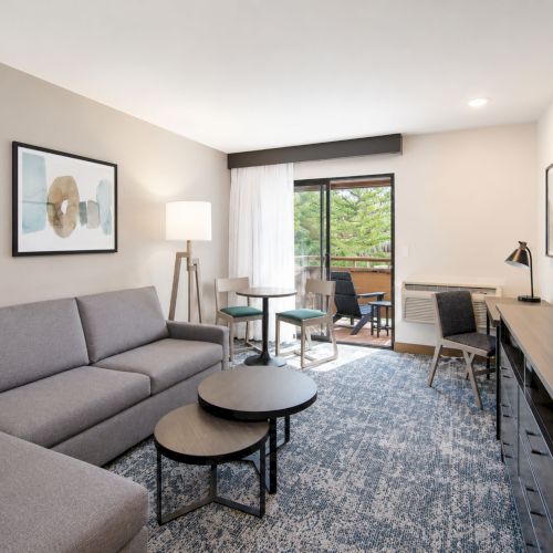 A modern living room with a gray sofa, coffee table, TV wall, dining chairs, and a sunny view through large windows.